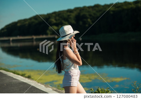 A young woman in white stands by the lake, immersed in conversation 127080483