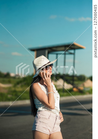 A girl wearing white shorts, a T-shirt and a hat is standing and holding the phone to her ear A girl wearing white shorts, a T-shirt and a hat is standing and holding the phone to her ear 127080485
