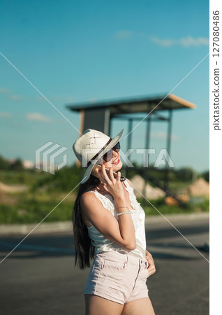 A young woman in a light white outfit is standing in the middle of nature and talking on the phone A young woman in a light white outfit is standing in the middle of nature and talking on the phone 127080486