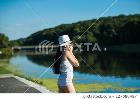 A slender girl by the water, dressed in a T-shirt and shorts, holds a phone and talks A slender girl by the water, dressed in a T-shirt and shorts, holds a phone and talks 127080487