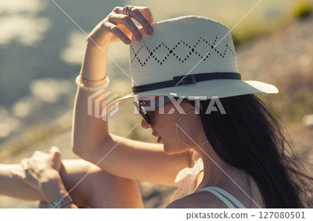 A girl wearing sunglasses and a hat is sitting by the lake, exposing her face to the sun's rays 127080501