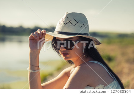 A girl wearing a hat and glasses sits near a quiet lake, basking in the rays 127080504