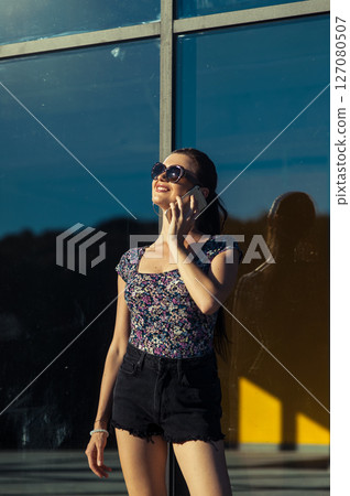 A girl wearing a light T-shirt and shorts is standing next to a glass building, holding a cell phone to her ear 127080507