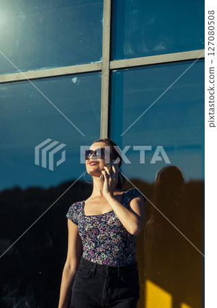 A young woman with glasses and a cell phone in her hands is having a conversation near a glass structure 127080508