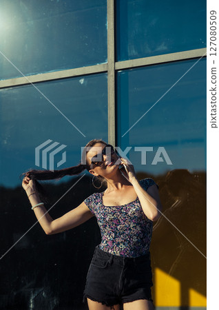 A young woman with glasses and a cell phone in her hands is having a conversation near a glass structure A young woman with glasses and a cell phone in her hands is having a conversation near a glass structure 127080509