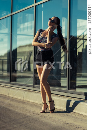 A young woman in a stylish outfit is talking on the phone next to a glass structure A young woman in a stylish outfit is talking on the phone next to a glass structure 127080514