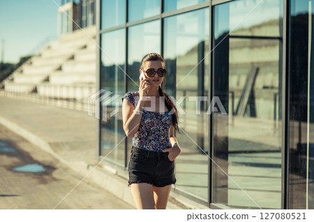 A young woman in shorts stands next to a shiny building, holding a phone A young woman in shorts stands next to a shiny building, holding a phone 127080521