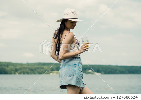 A young girl stands by the lake with a bright cup of juice in her hand A young girl stands by the lake with a bright cup of juice in her hand 127080524