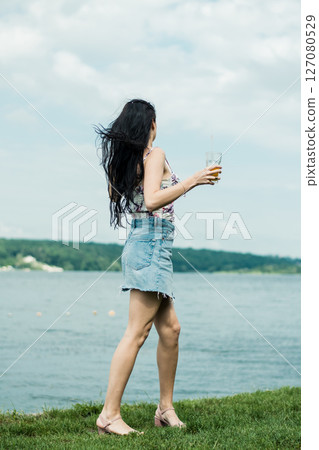 The girl in a hat has stopped by the lake, enjoying the drink in her cup The girl in a hat has stopped by the lake, enjoying the drink in her cup 127080529