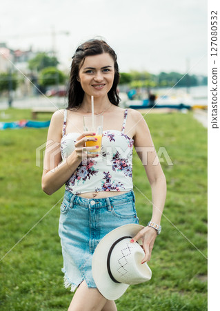A young girl stands by the lake with a glass of juice in her hands 127080532