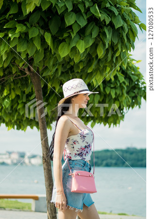 A young girl in a skirt, tank top, and hat stands by the lake with a pink handbag on her shoulder 127080533