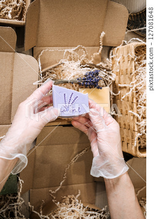 Gloved hands hold lavender soap above shredded filler and open kraft packaging. Product presentation, boutique branding, handmade business, artisan soap design 127081648