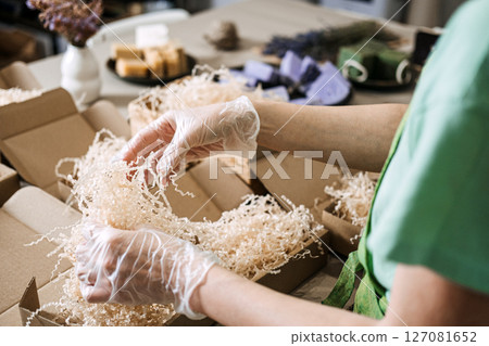 Gloved hands arrange green and lavender soaps with dried flowers in kraft packaging. Handmade business, eco-friendly packaging, natural skincare, artisan presentation 127081652