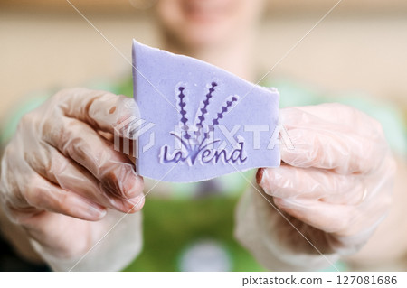 Smiling person in gloves holds up a stamped lavender soap bar for the camera. Lavender soap, handmade branding, aromatherapeutic skincare, plant-based wellness 127081686