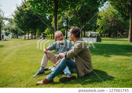 Father and adult son sitting on grass in park, sharing a story from the past. 127081745