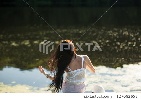 A young lady in light summer clothes is sitting by the lake water, taking a break from the city noise A young lady in light summer clothes is sitting by the lake water, taking a break from the city noise 127082655