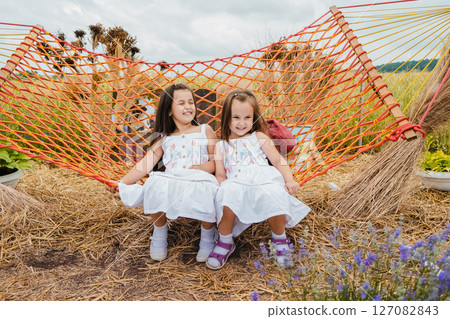 Two girls in white dresses are joyfully swinging on a red mesh hammock Two girls in white dresses are joyfully swinging on a red mesh hammock 127082843