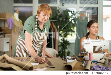 Mature dressmaker drawing patterns next to young assistant 127086357
