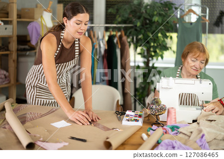 Young Dressmaker Drawing on Paper Next to Mature Female Seamstress Young Dressmaker Drawing on Paper Next to Mature Female Seamstress 127086465