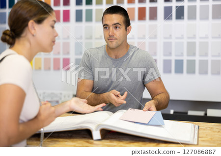 Male salesman of paintwork material shop advises woman buyer with samples and product catalog 127086887