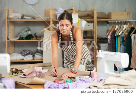 Female dressmaker draws sketch of future clothing on paper using patterns in sewing workshop Female dressmaker draws sketch of future clothing on paper using patterns in sewing workshop 127086900