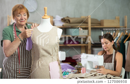 Elderly dressmaker stands near a mannequin and checks the dress against the background of the assistant 127086938