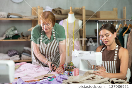 Mature woman cutting fabric next to female seamstress Mature woman cutting fabric next to female seamstress 127086951