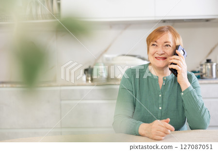 Elderly positive woman stands at home in kitchen and talks nicely on phone with friend 127087051