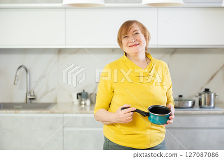 Elderly woman in casual clothes stands in front of table and holds coffee pot Elderly woman in casual clothes stands in front of table and holds coffee pot 127087086