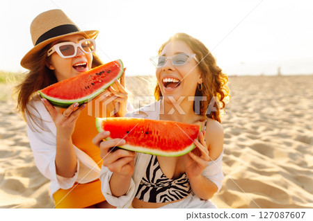 Smiling two women eating watermelon on beach. Happy summer time. People, lifestyle, travel Smiling two women eating watermelon on beach. Happy summer time. People, lifestyle, travel 127087607