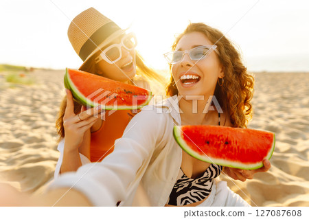Smiling two women eating watermelon on beach. Happy summer time. People, lifestyle, travel Smiling two women eating watermelon on beach. Happy summer time. People, lifestyle, travel 127087608