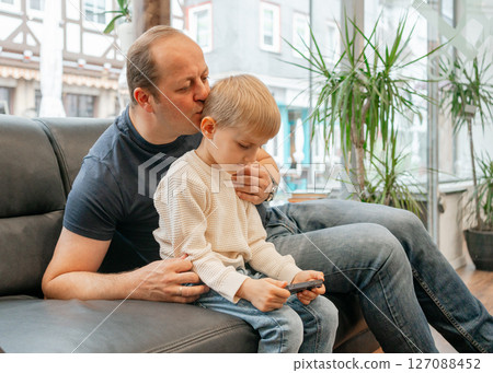 Father lovingly kisses son on the head while he plays on a phone, sitting together on a comfortable couch. The scene captures a warm moment of affection and bonding. 127088452