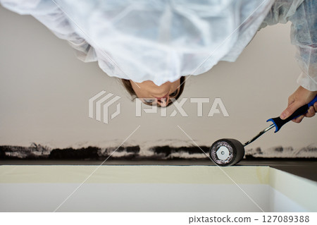 Woman in protective suit painting wall and ceiling with paint roller in black color during interior renovation Woman in protective suit painting wall and ceiling with paint roller in black color during interior renovation 127089388