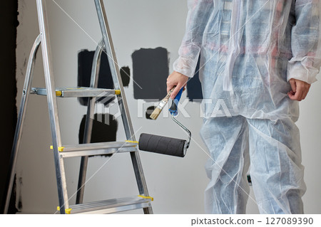 Close-up view of person in protective coveralls holding paint roller and brush near ladder. Wall painting process during home renovation Close-up view of person in protective coveralls holding paint roller and brush near ladder. Wall painting process during home renovation 127089390