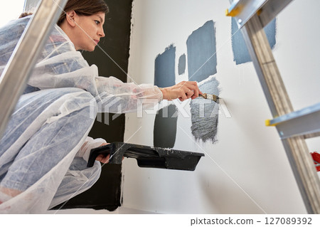 Woman in protective wear applies different dark paint swatches on white wall using brush. Female painter testing black and dark gray paint samples during interior renovation project in modern home Woman in protective wear applies different dark paint swatches on white wall using brush. Female painter testing black and dark gray paint samples during interior renovation project in modern home 127089392