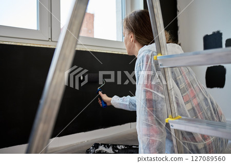 Woman in protective suit stands on ladder and paints wall black with roller in room under renovation. Interior painting process during home improvement Woman in protective suit stands on ladder and paints wall black with roller in room under renovation. Interior painting process during home improvement 127089560