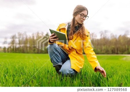 Young farmer woman in a green field, intently using a tablet. She wears a yellow jacket and glasses 127089568