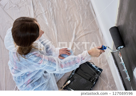 Woman in protective suit painting interior wall black with roller brush in modern room under renovation Woman in protective suit painting interior wall black with roller brush in modern room under renovation 127089596