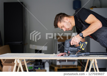 Worker in workwear using handheld cordless electric jigsaw to cut wooden board in garage. Skilled carpenter working in workshop. Man doing DIY home renovation 127089607