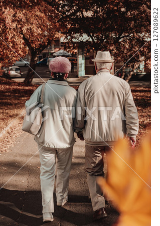 Elderly couple walking in autumn park. Elderly couple in light jackets walks together through peaceful autumn park with golden foliage. Concept of companionship, aging with love, autumn stroll Elderly couple walking in autumn park. Elderly couple in light jackets walks together through peaceful autumn park with golden foliage. Concept of companionship, aging with love, autumn stroll 127089622