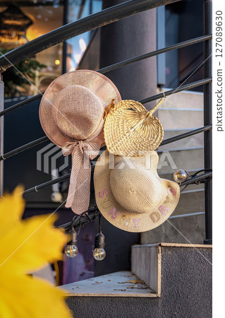 Straw hats hanging on railing. Three straw hats in different styles hang on metal railing outside near entrance, warm sunlight. Concept of summer end, change of seasons, arrival of autumn, nostalgia Straw hats hanging on railing. Three straw hats in different styles hang on metal railing outside near entrance, warm sunlight. Concept of summer end, change of seasons, arrival of autumn, nostalgia 127089630