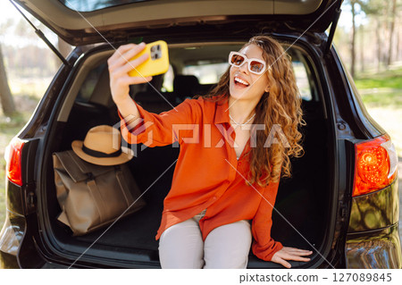 Happy woman sits on the trunk of her car and taking a selfie. Selfie while traveling by car. Happy woman sits on the trunk of her car and taking a selfie. Selfie while traveling by car. 127089845