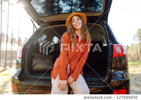 A traveler young woman smiles while sitting in the trunk of her car during a sunny day in a forest 127089850