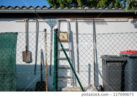 Garden tools and ladder hanging on white wall behind fence, concept of outdoor organization Garden tools and ladder hanging on white wall behind fence, concept of outdoor organization 127090673