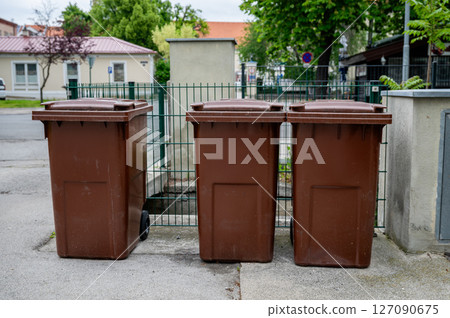 Three brown garbage bins aligned on street near fence and residential buildings. Three brown garbage bins aligned on street near fence and residential buildings. 127090675