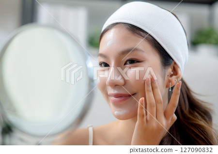 Beauty young asian woman applying cream on facial for wrinkles while looking mirror in living room. Beauty young asian woman applying cream on facial for wrinkles while looking mirror in living room. 127090832