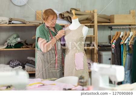 Mature female tailor sews new dress on a mannequin in sewing studio 127090869