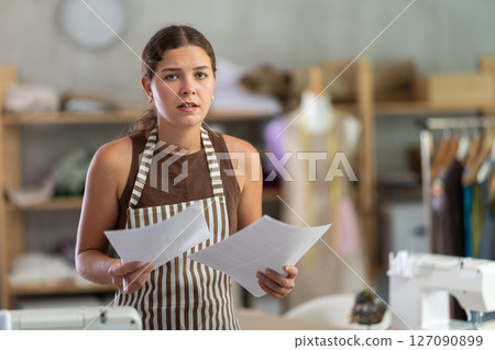 Sad seamstress girl reading financial document in sewing studio interior Sad seamstress girl reading financial document in sewing studio interior 127090899