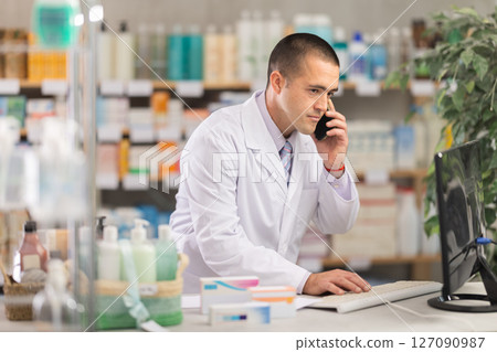 Man pharmacist works at computer and talks on phone 127090987