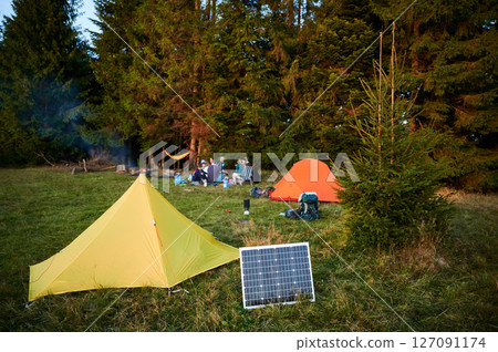 Solar panel in grassy field with tents in background highlights eco-friendly camping setup. Scene includes campers, emphasizing integration of renewable energy in outdoor activities. Solar panel in grassy field with tents in background highlights eco-friendly camping setup. Scene includes campers, emphasizing integration of renewable energy in outdoor activities. 127091174